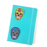 Twee strijk patches van een rijkversierde paars / lila en gele sugar skull  Met geborduurde versiering accenten op de voorzijde van een blauw / groen notietieboekje